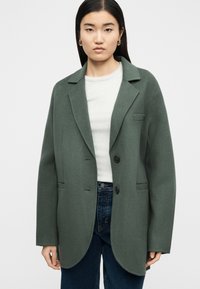 Γυναίκα φορώντας υπερμεγέθη πράσινο μάλλινο σακάκι πάνω από λευκή ριμπ τοπ και μπλε τζιν, κοιτάζοντας μπροστά με ουδέτερη έκφραση σε απλό φόντο.