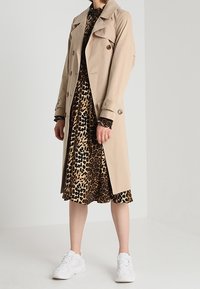 Trench coat beige con design doppiopetto e colletto, indossato sopra un vestito con stampa leopardata, abbinato a scarpe da ginnastica bianche.