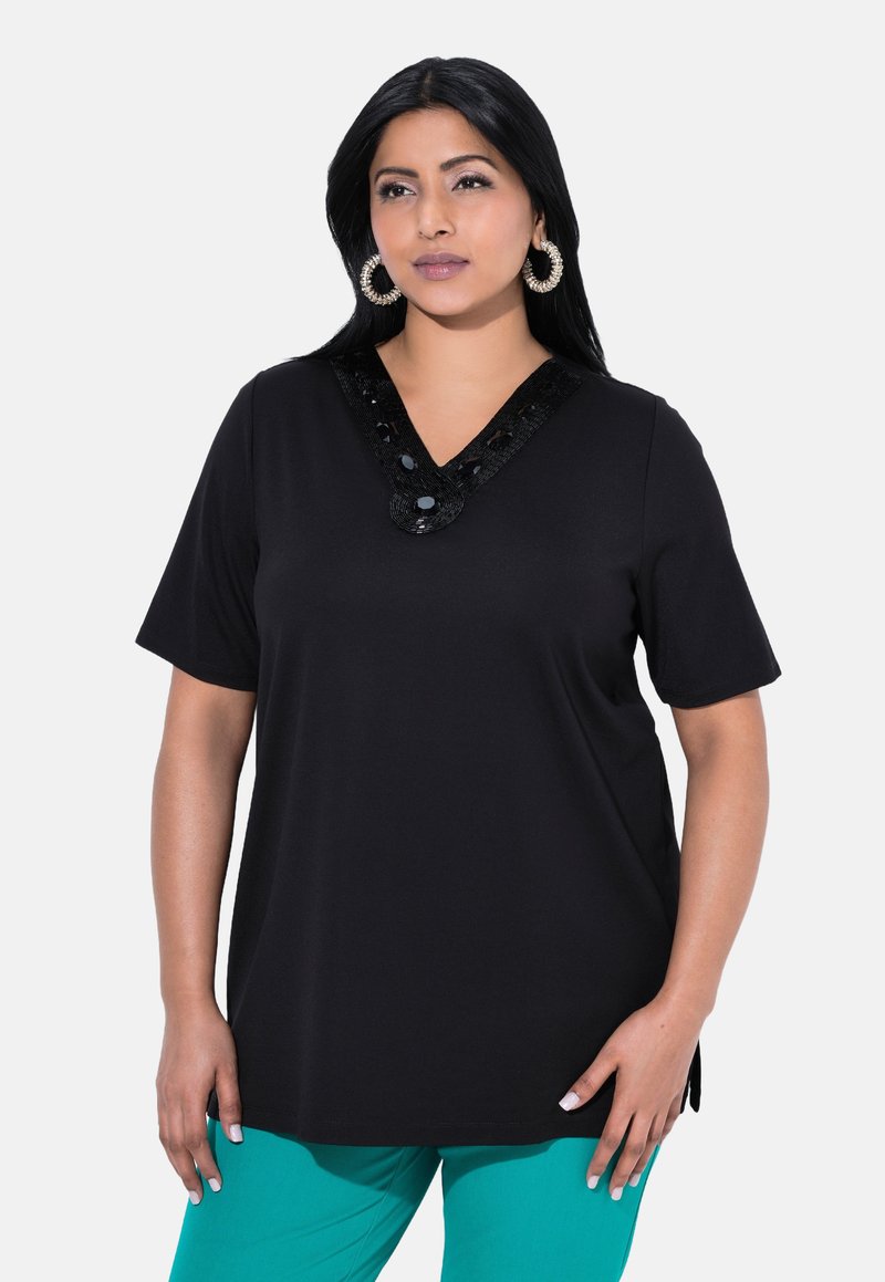 Camiseta negra de cuello en V con mangas cortas, que presenta detalles de lentejuelas en el escote, combinada con pantalones en color verde azulado. Textura suave, corte relajado.