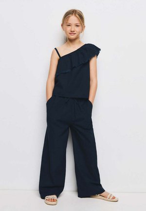 Jeune fille debout contre un mur blanc, portant un top asymétrique à volants bleu marine, un pantalon large et des sandales blanches avec des brides tressées.