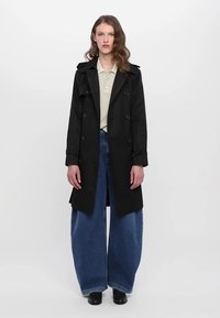 Casaco trench preto com botões duplos, lapelas largas e punhos ajustáveis, usado sobre uma camisa polo às riscas bege e jeans de perna larga azuis.