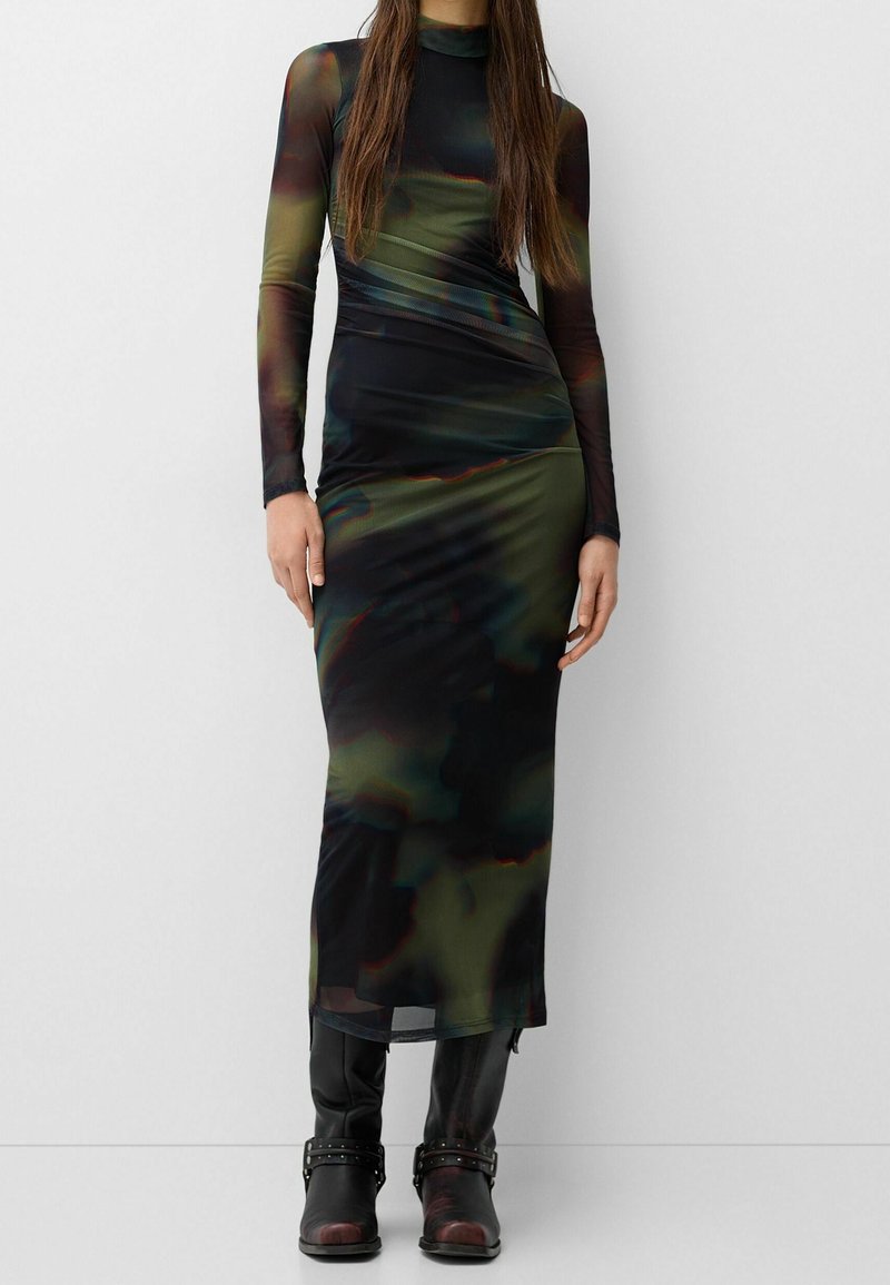 Femme portant une robe maxi moulante à manches longues et col montant avec un motif abstrait sombre, associée à des bottines noires à brides.