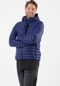 Marineblauwe gewatteerde jas met een capuchon, featuring een ritssluiting aan de voorkant en zijzakken. Het gestikte ontwerp voegt textuur toe. Draagt over zwarte broek.
