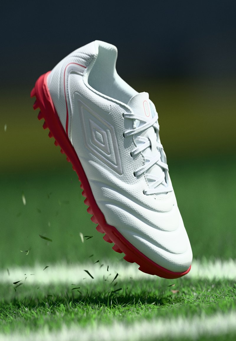 Zapatilla deportiva blanca con una superficie texturizada, suela roja y cordones, que presenta un detalle del logo de diamante. Situada sobre hierba con escombros esparcidos.