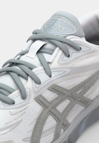 Vitaid sportskor i vitt med grå detaljer, texturerat mesh-överkott, grå snören och en framträdande Asics-logotyp på tungan.
