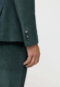 Sötétzöld kordbársony blézer ujja, amely texturált bordázott mintázattal rendelkezik, és négy fekete gomb található a mandzsettáján.