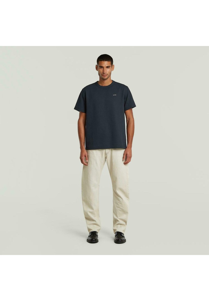 Hombre de pie contra un fondo liso, vistiendo una camiseta azul marino oscuro, pantalones beige claros y zapatos negros, mirando hacia adelante con las manos relajadas.