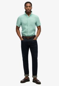 Koszulka z krótkim rękawem w miętowym kolorze z lnu, ciemnoniebieskie dżinsy z podwiniętymi mankietami, brązowy skórzany pasek i ciemnobrązowe buty z fakturowaną powierzchnią.