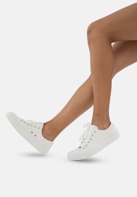 Białe płócienne trampki z zaokrąglonym noskiem, sznurowane z przodu, z metalowymi oczkami i teksturowaną gumową podeszwą. Wyeksponowane na skośnej nodze.