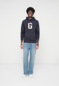 Marine sweatshirt med et cremefarvet logo, frontlomme og hætte med snor; parret med lyseblå lige bukser og hvide sneakers.