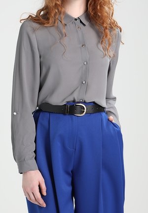 Kvinde iført en grå skjorte med knapper, stukket ned i højtaljede blå bukser med sort bælte, hånd i lommen, foran en ensfarvet baggrund.