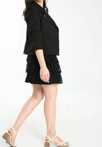 Veste noire courte avec un design à col, associée à une jupe noire à volants avec des détails en dentelle et des sandales blanches à talons bloc.