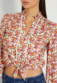 Bloemige blouse van witte katoen met roze, oranje en groene patronen. Heeft een strik in de taille, een mandarinenkraag en lange mouwen.