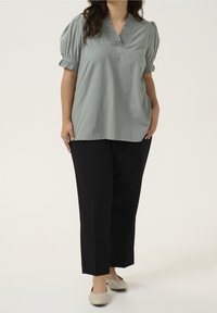 Vrouw in een saliegroene blouse met pofmouwen, zwarte recht-pas pantalon en beige ballerina's, staand tegen een effen achtergrond.