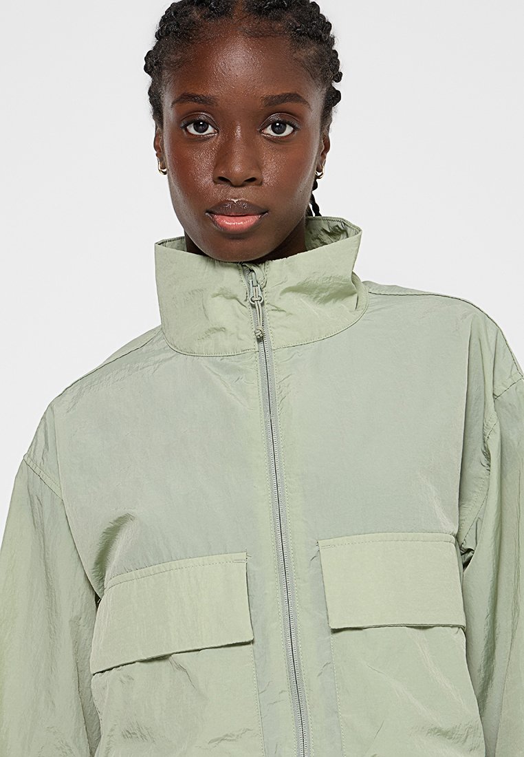 Veste zippée vert clair avec un col haut, deux poches plaquées et un tissu texturé. Design respirant et décontracté, idéal pour le superposition.