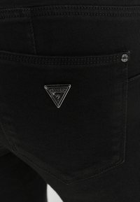 Jeans negros con un bolsillo trasero que presenta un logo triangular plateado de Guess y un ribete de contraste, destacando un tejido suave y texturizado.