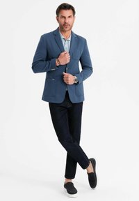 Man i blå kavaj, ljusblå polo, mörka jeans, svarta slip-on skor, står med benen korsade mot vit bakgrund.