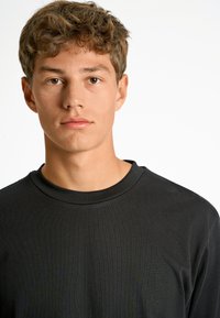 T-shirt nera in cotone con collo rotondo; texture morbida e vestibilità rilassata. Il viso del modello è centrato, con pelle chiara e capelli corti e disordinati.