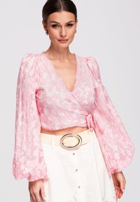 Femme portant un chemisier cache-cœur rose à fleurs avec des manches longues bouffantes et une jupe blanche taille haute avec une grande boucle de ceinture ovale.