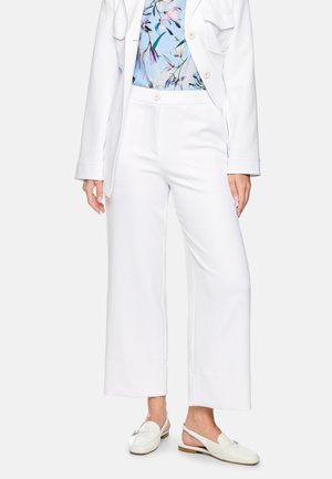 Woman wearing white wide-leg pants, white blazer, blue floral top, and white slingback loafers standing against plain background.