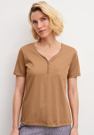 Vrouw met kort blond haar die een beige korte mouw henley draagt en paarse broek met sterpatroon, staand tegen een witte achtergrond.
