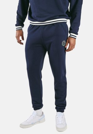 Man draagt marineblauwe joggers en een bijpassend sweatshirt met witte en zwarte gestreepte boorden, gecombineerd met witte sneakers en sokken.