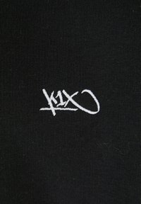 Tessuto nero con una trama liscia, caratterizzato da un logo ricamato bianco in forma di scrittura stilizzata, posizionato centralmente sul tessuto.