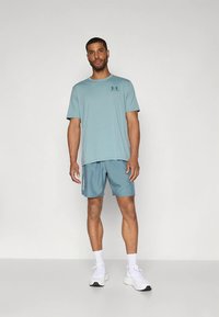 Mees seisab, kandes helesinist Under Armour t-särki ja sobivaid lühikesi pükse, valgeid sokke ning valgeid tossusid lihtsal valgel taustal.