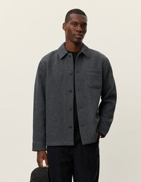 Grijze herringbone overhemdjas met een kraag, knoopsluiting aan de voorkant en borstzak. Gemaakt van textuurstof, gecombineerd met donkere broeken.