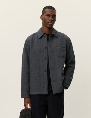 Grijze herringbone overhemdjas met een kraag, knoopsluiting aan de voorkant en borstzak. Gemaakt van textuurstof, gecombineerd met donkere broeken.