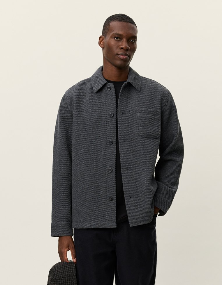Grijze herringbone overhemdjas met een kraag, knoopsluiting aan de voorkant en borstzak. Gemaakt van textuurstof, gecombineerd met donkere broeken.