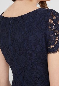Robe en dentelle bleu marine avec fermeture éclair dissimulée dans le dos et manches courtes en dentelle affichant des motifs floraux et un bord festonné.