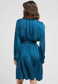 Robe teal avec des manches longues, une taille smockée et un tissu fluide qui arrive au-dessus du genou, avec une finition texturée subtile.