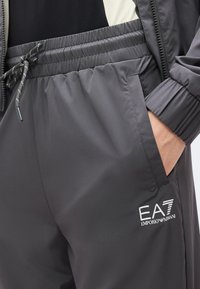 Κοντινό πλάνο ατόμου που φοράει σκούρο γκρι παντελόνι φόρμας Emporio Armani EA7 με ελαστική μέση και κορδόνι, χέρι στην πλαϊνή τσέπη.