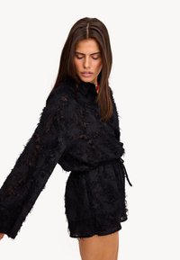 Robe chemise noire transparente avec un tissu texturé et effiloché. Présente des manches longues, un devant à boutons et une ceinture à la taille pour un ajustement réglable.