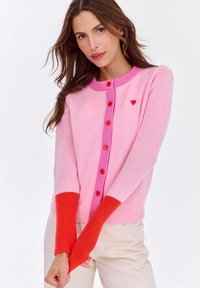 Różowy i czerwony sweter z dzianiny z długimi, prążkowanymi czerwonymi rękawami, guzikami z przodu oraz małym czerwonym serduszkiem przy dekolcie.