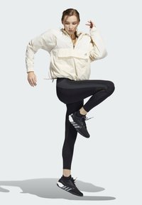 Beige leichte Jacke mit einer Fronttasche, schwarze hochgeschnittene Leggings und schwarze Sportschuhe mit weißen Streifen, in einer dynamischen Pose.