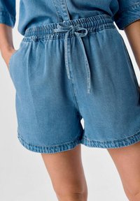 Shorts de mezclilla en azul claro, con una cinturilla elástica y cordón ajustable, además de bolsillos laterales. El dobladillo está vuelto y cosido.