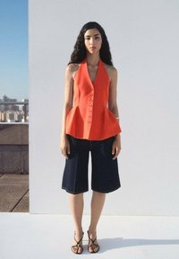 Orange peplum top with button detail, paired with dark denim culottes and black strappy sandals. Model stands against a light background.