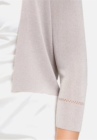 Ljusbeige cardigan med trekvartsärmar. Har en texturerad stickning och små dekorativa hål vid ärmsluten.