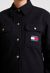 Ciemnoniebieska koszula z guzikami, wykonana z teksturowanej tkaniny, z dwoma kieszeniami na piersi oraz małą naszywką z logo w czerwonym, białym i niebieskim kolorze.