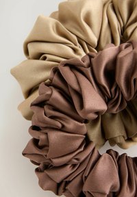 Kaks kangast scrunchy-d; üks beež ja teine pruun. Plekkide tekstuur, pehme materjal ja elastne disain, pildil veidi kattuvad.