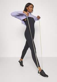 Body in tuta manica lunga nera e maglione viola chiaro. La modella tira una banda elastica indossando scarpe da ginnastica nere. Sfondo semplice.