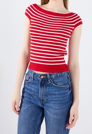 Frau trägt ein rot-weiß gestreiftes ärmelloses Oberteil mit horizontalen Streifen, kombiniert mit hoch taillierten blauen Jeans, steht vor einem schlichten Hintergrund.