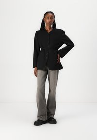 Veste noire cintrée avec un col en V, devant à bouton et taille ceinturée, associée à un jean large gris clair et des chaussures noires à semelle épaisse.