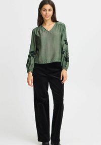 Donna che indossa una blusa verde a maniche lunghe con motivi di foglie nere sulle maniche e pantaloni larghi neri, in piedi davanti a uno sfondo semplice.