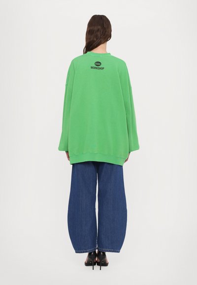 Grønn oversized genser med ribbestrikkede mansjetter og falden, med en svart logo på baksiden. Kombinert med brede blå denimbukser.