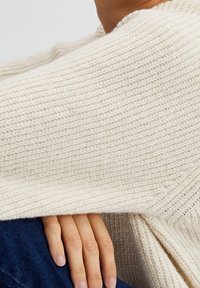 Maglione spesso in crema lavorato a maglia con un motivo testurizzato. L'orlo è leggermente sciolto, mostrando una manica che pende su una mano.