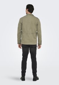 Giacca verde oliva con vita regolabile, spalline e polsini con bottoni. Abbinata a jeans neri a taglio slim e stivali neri.