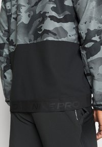 Sportovní top Nike Pro s šedým maskáčovým designem, černým dolním panelem a strukturovanou tkaninou. Obsahuje logo na lemování a přiléhavý siluetu.